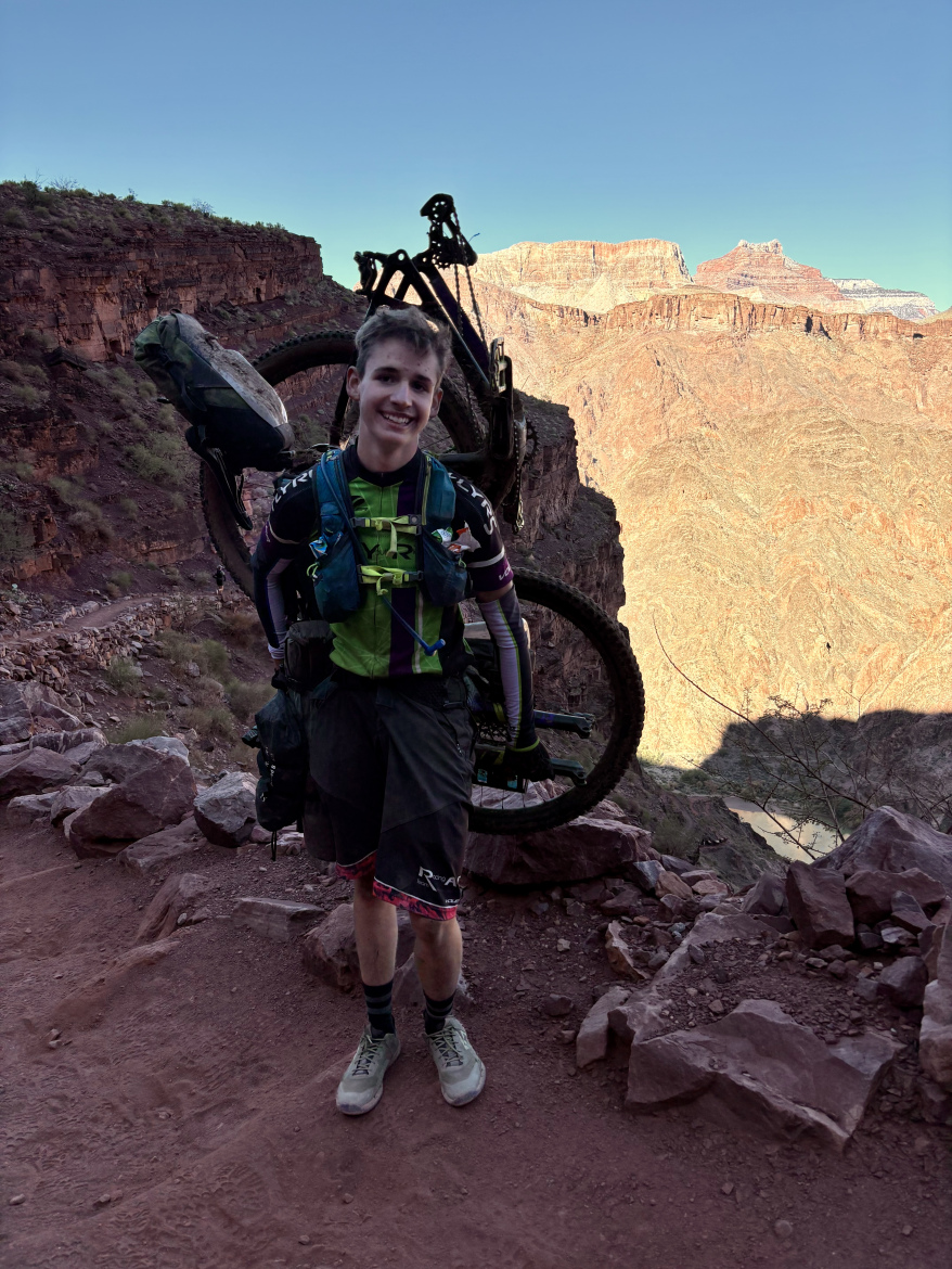 A young man stands on a rocky trail, holding a mountain bike over his shoulder. He is wearing a bright green and black athletic outfit, along with gray shoes and colorful socks. The background features a stunning view of steep canyon walls and a clear blue sky. The terrain appears rugged, suggesting an adventurous outdoor activity.