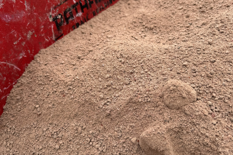 A close-up view of a wheelbarrow filled with fine, sandy material, featuring a worn red surface and a visible logo. The texture of the sand appears smooth, with some small clumps.