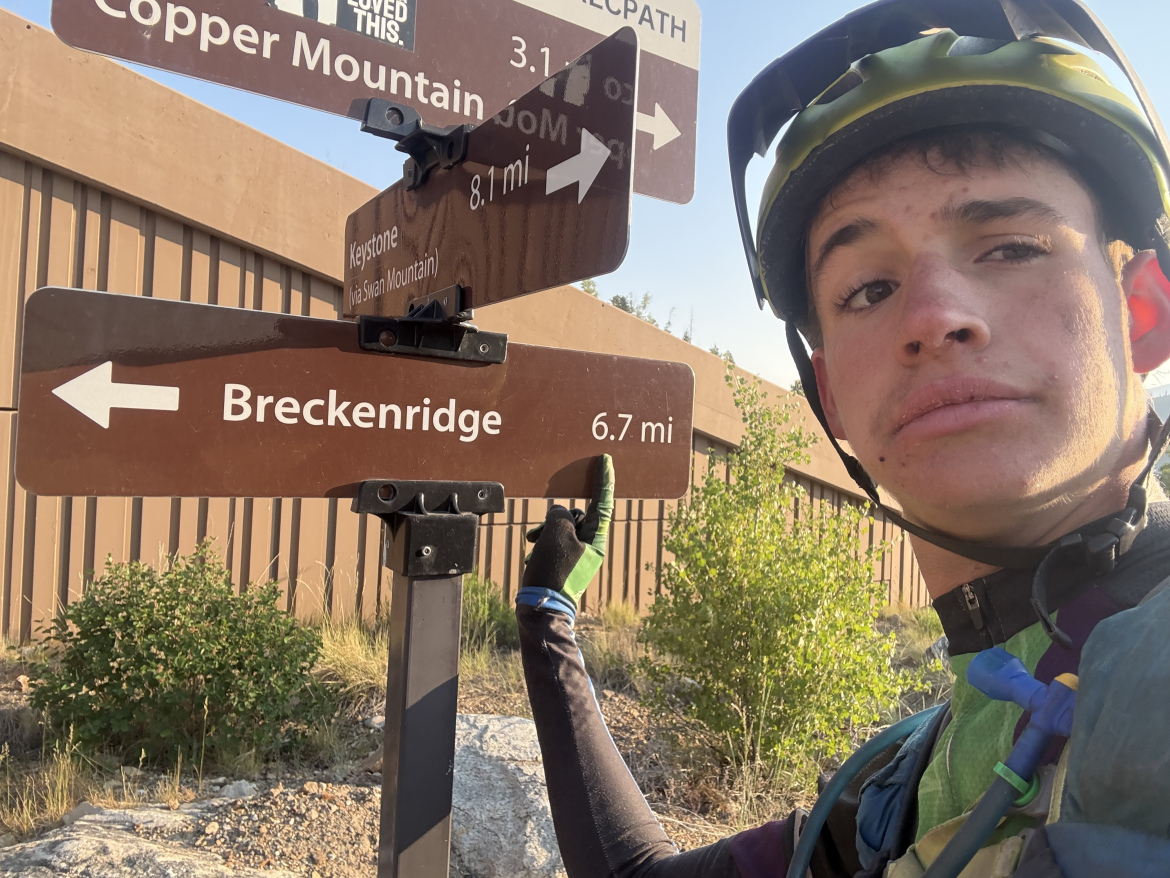 A young person wearing a cycling helmet and gear stands next to a trail sign indicating directions to Breckenridge, Copper Mountain, and Keystone. The sign shows the distance to Breckenridge is 6.7 miles. In the background, there are trees and a brown fence, with a clear blue sky above.
