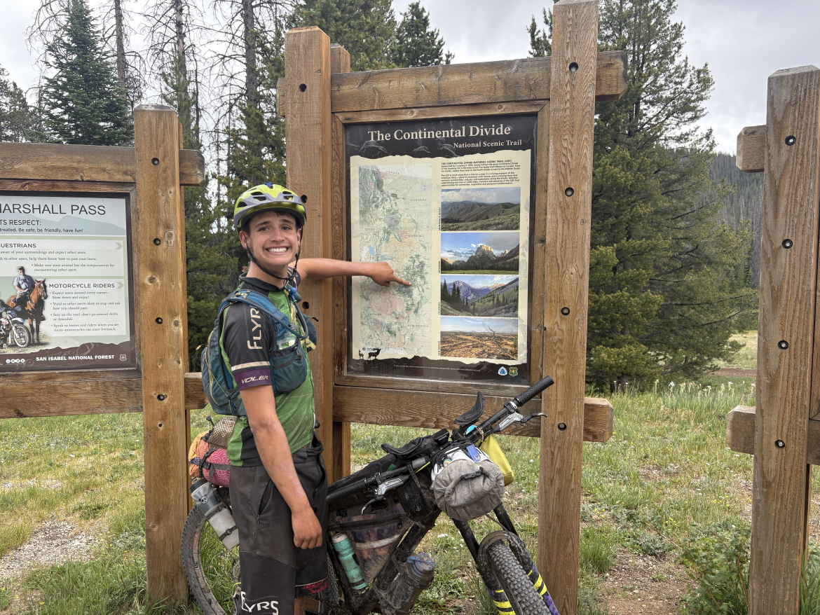 A young man wearing a helmet and a cycling vest smiles and points at a map displayed on a wooden sign at a trailhead. The map is labeled "The Continental Divide National Scenic Trail" and shows scenic images of landscapes. The man is standing next to his bike, which is loaded with gear, surrounded by tall trees and grass in a mountainous area.