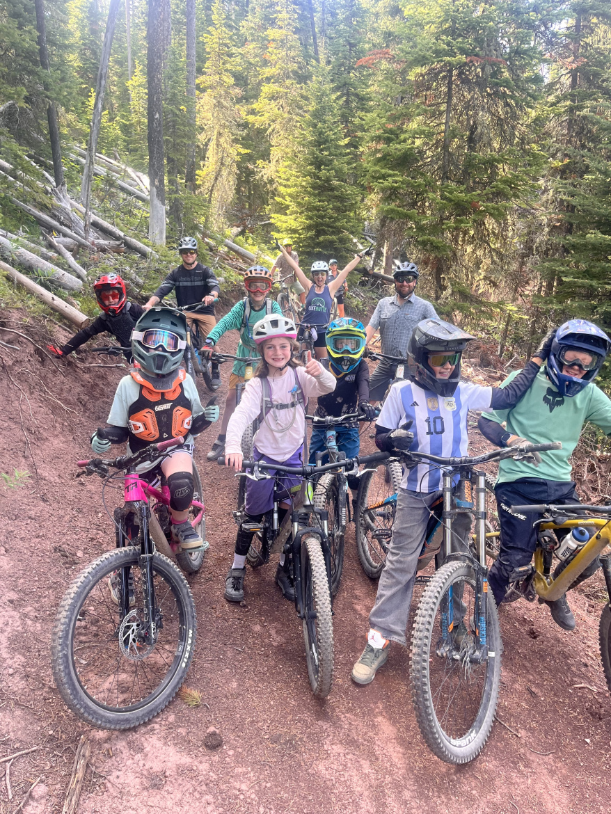 A group of children and adults are posing for a photo on mountain bikes in a forested area. They are wearing helmets and protective gear, with a variety of colorful outfits. The background features tall trees and a dirt path, creating a vibrant natural setting. The group is smiling and showing signs of excitement.