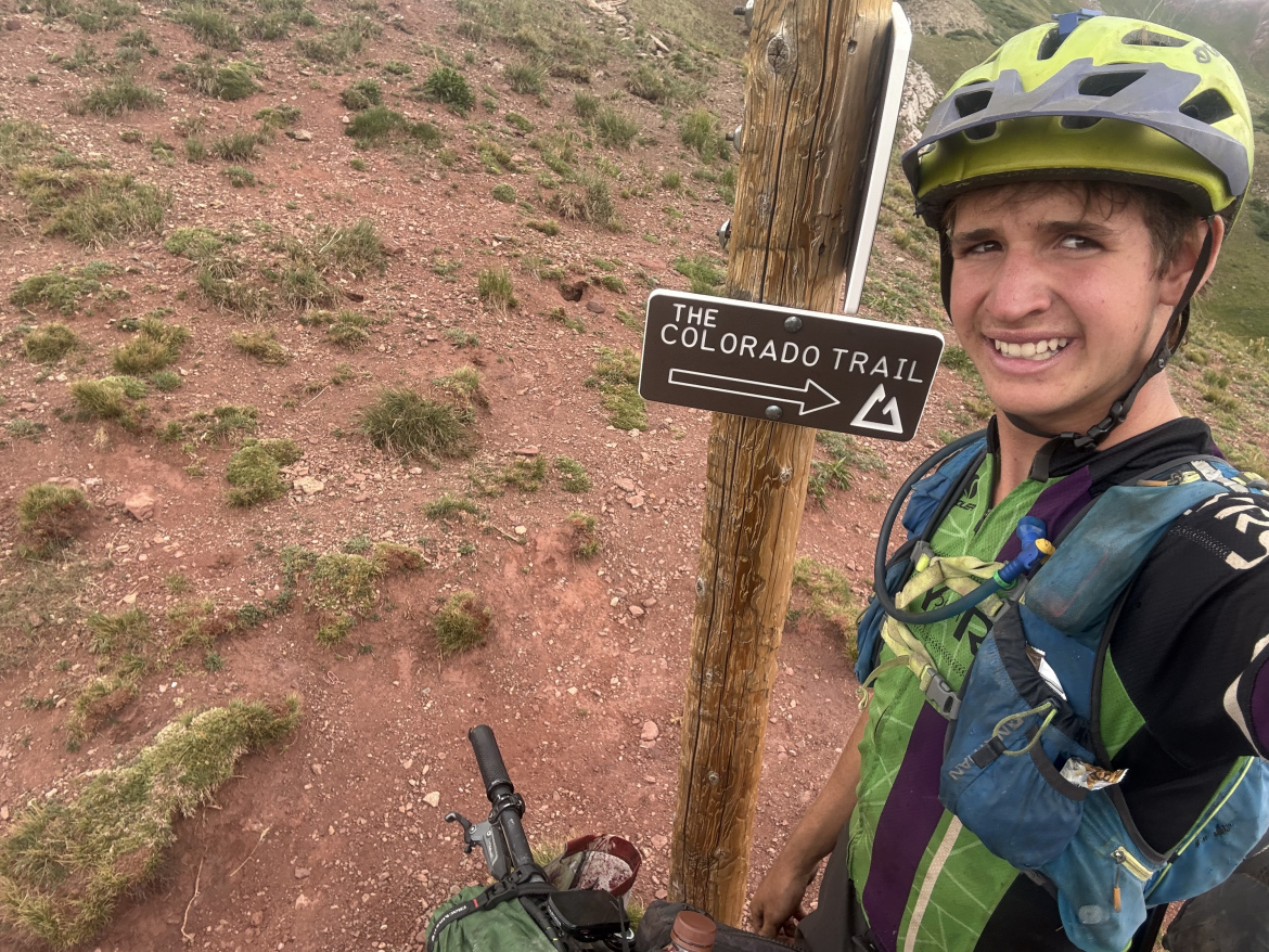 A person wearing a helmet and cycling gear stands next to a directional sign that indicates the Colorado Trail, with a landscape of grassy and rocky terrain in the background. The individual is holding their bike, wearing a backpack, and smiling for the photo.