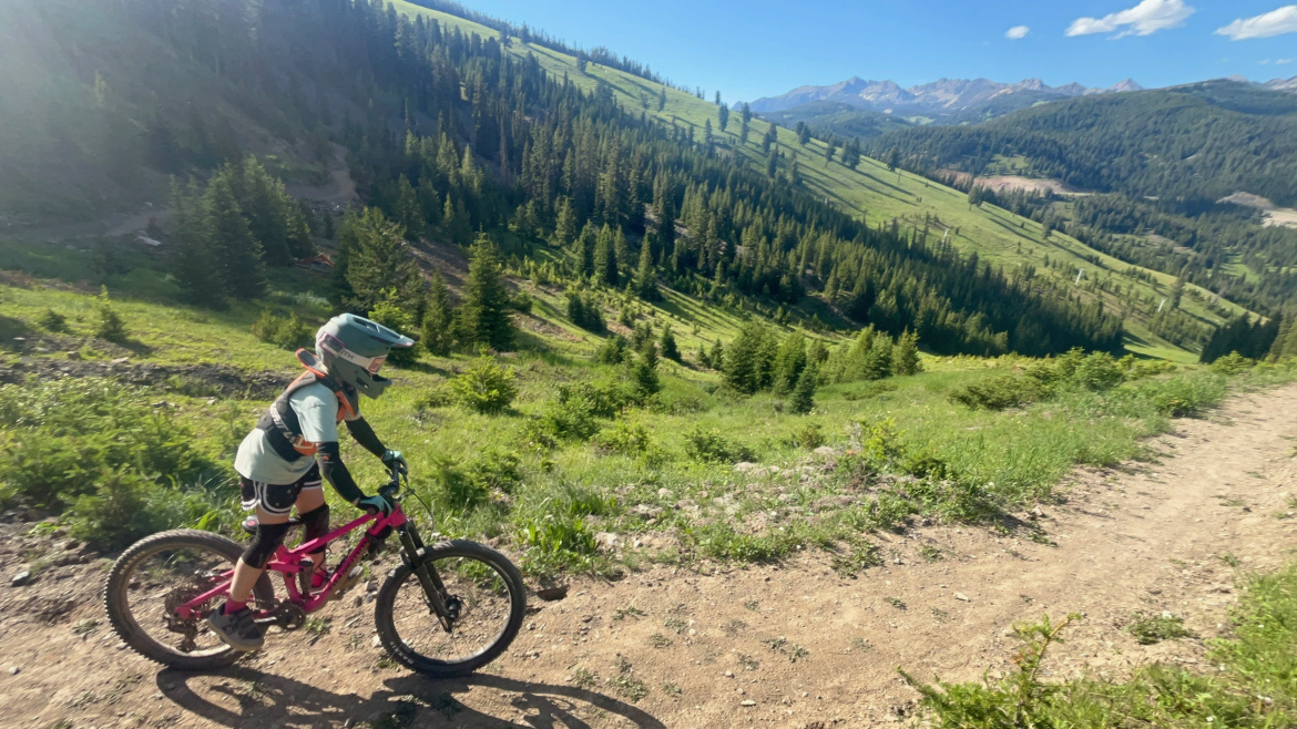 A child in protective gear rides a bright pink mountain bike along a dirt trail in a lush green landscape, with a backdrop of towering trees and distant mountains under a clear blue sky.
