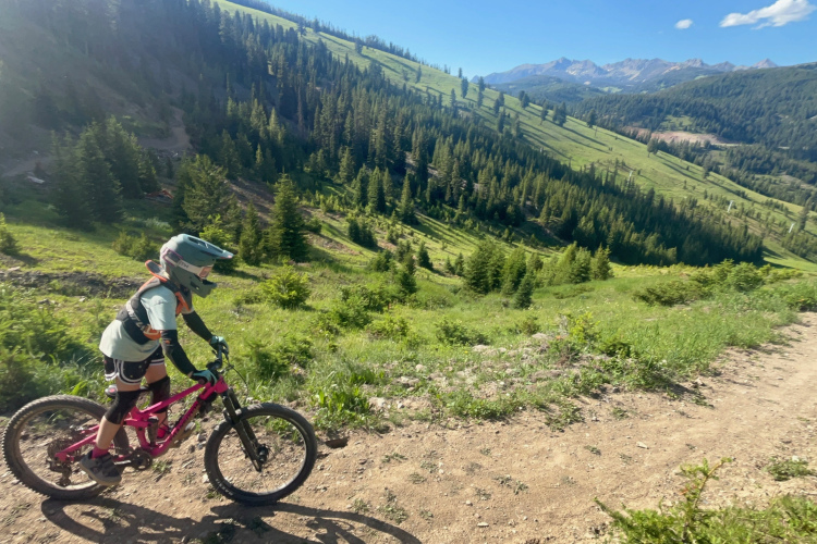 A child in protective gear rides a bright pink mountain bike along a dirt trail in a lush green landscape, with a backdrop of towering trees and distant mountains under a clear blue sky.
