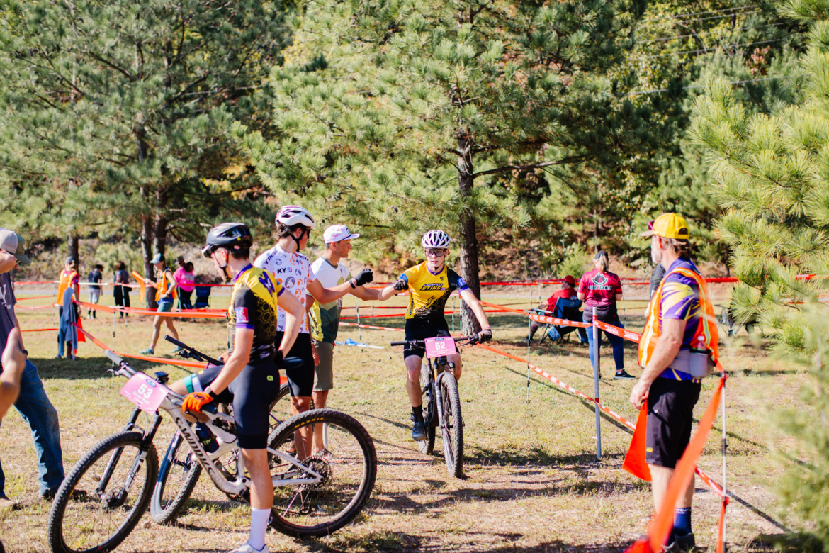 A mountain biking event scene showing several competitors in jerseys with their bicycles. Two cyclists are interacting, with one cyclist receiving a fist bump from a supporter. In the background, spectators are seated, and there are trees and event barriers nearby. The atmosphere is lively and supportive, typical of a sports competition.