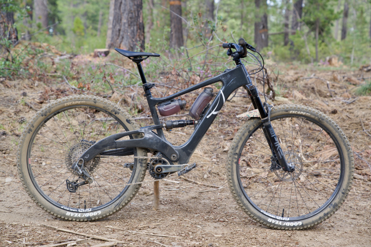 A mountain bike with a black frame and distinct suspension design is positioned on a dirt trail surrounded by trees. The bike features wide tires with visible tread, a sleek seat, and a pair of water bottles mounted in the frame. The background consists of a natural wooded area with scattered leaves and small branches.