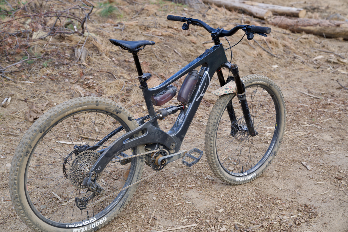 A black mountain bike resting on a dirt trail, featuring large, knobby tires and a sleek frame. The bike has a visible rear suspension system, and two water bottle holders are mounted on the frame. Surrounding the bike is a natural setting with dried leaves and scattered branches.