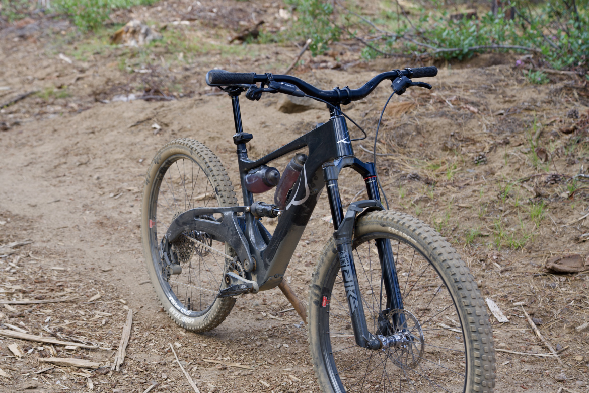 A close-up view of a mountain bike positioned on a dirt trail, with visible tire tread and a water bottle mounted on the frame. The bike has a sleek design and is surrounded by a natural setting with dirt and foliage in the background.