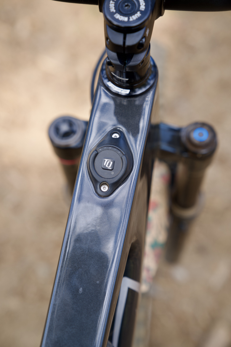 Close-up view of a mountain bike frame featuring a black control button labeled "TQ," located on the top tube. The background shows a blurred natural terrain, likely dirt, suggesting an outdoor setting.