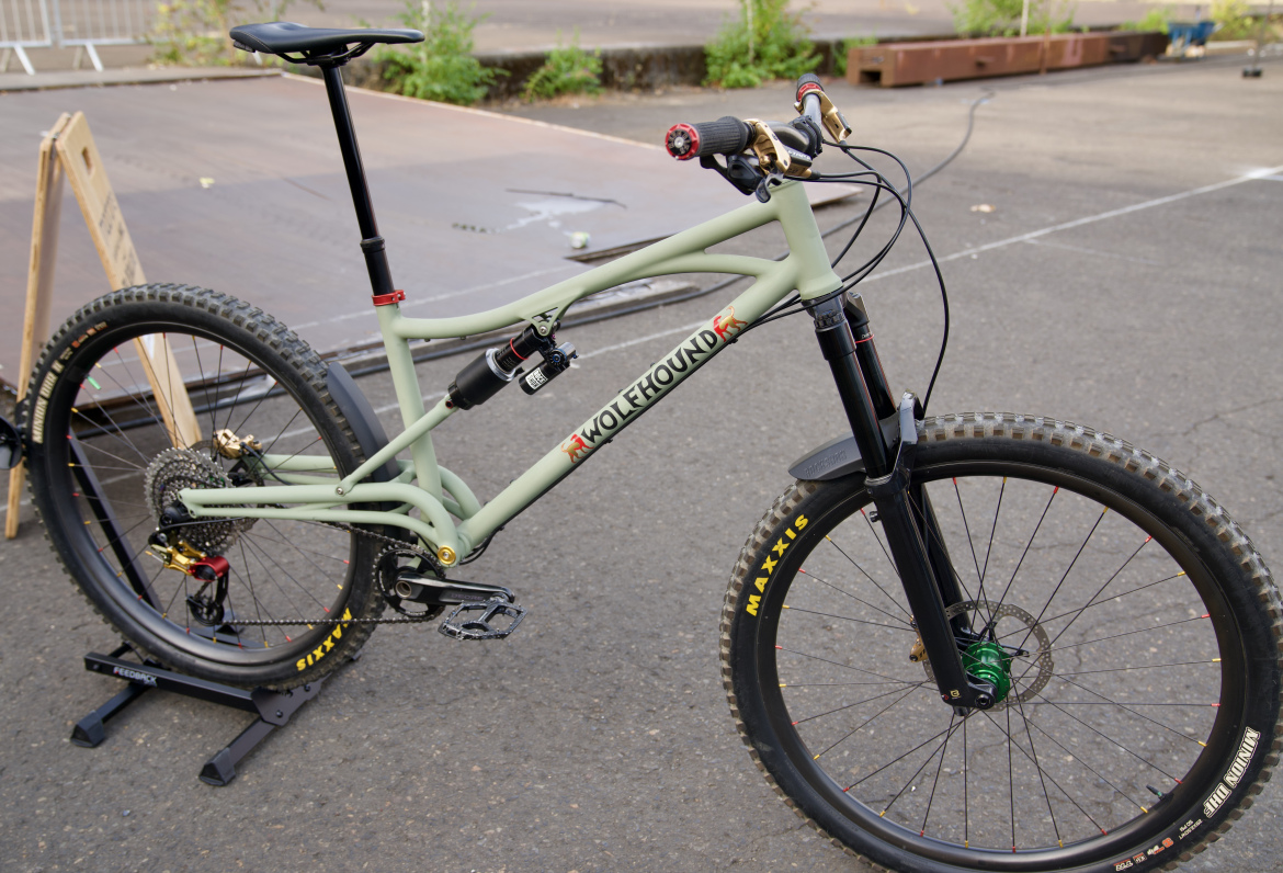 A close-up view of a mountain bike in a muted green color, showcasing its frame, suspension system, and components. The bike features thick tires with a tread pattern suitable for rough terrain, a dropper seat post, and disc brakes. The background includes a blurred outdoor setting with materials for a biking event.