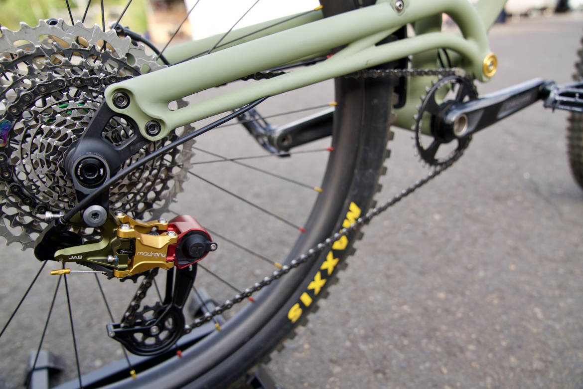 Close-up view of a mountain bike's rear drivetrain, featuring a green frame, gold and red derailleur, and a detailed cassette with visible chain links. The background shows a smooth, paved surface.