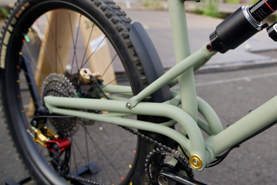 Close-up view of a bicycle's rear frame, showcasing the green matte finish, intricate linkages, and components including a rear shock, chain, and cogset. The background features a blurred outdoor setting with a hint of packaging materials.