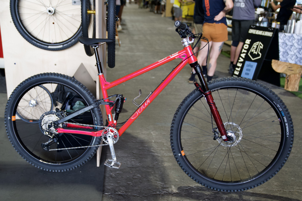 A bright pink mountain bike with a sleek frame and thick tires, displayed in a biking event setting. The bike features a suspension fork and a modern drivetrain, with a backdrop of other bicycles and event participants in a spacious indoor area.