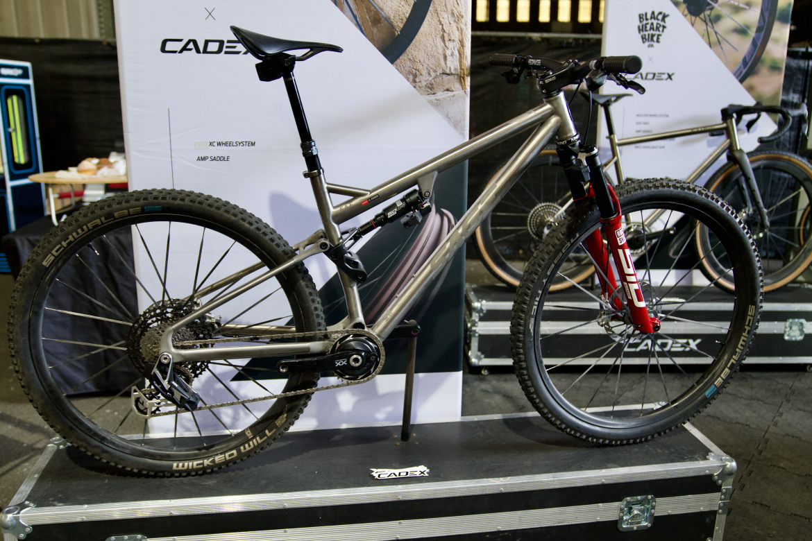 A modern mountain bike on a display stand, featuring a lightweight metallic frame, thick tires, and a striking red front suspension fork. The backdrop includes promotional banners for CADEX, showcasing bicycle components and accessories. The bike is equipped with a suspension system and gearing suitable for off-road riding.