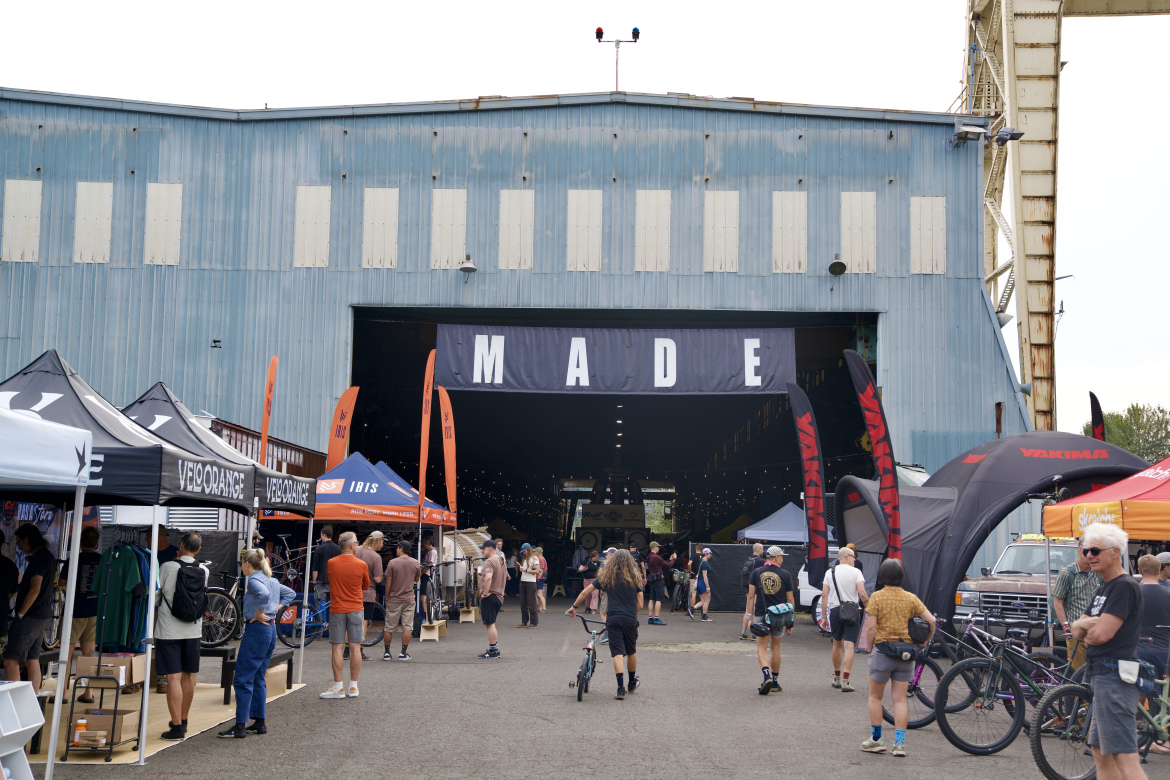 A large warehouse building with a blue exterior and banners reading "MADE" hangs above the entrance. Outside, various tents are set up displaying bicycles and cycling gear, and attendees are walking around, some on foot and others on bicycles. The atmosphere is lively, with people engaged in conversation and browsing the displays, under a cloudy sky.