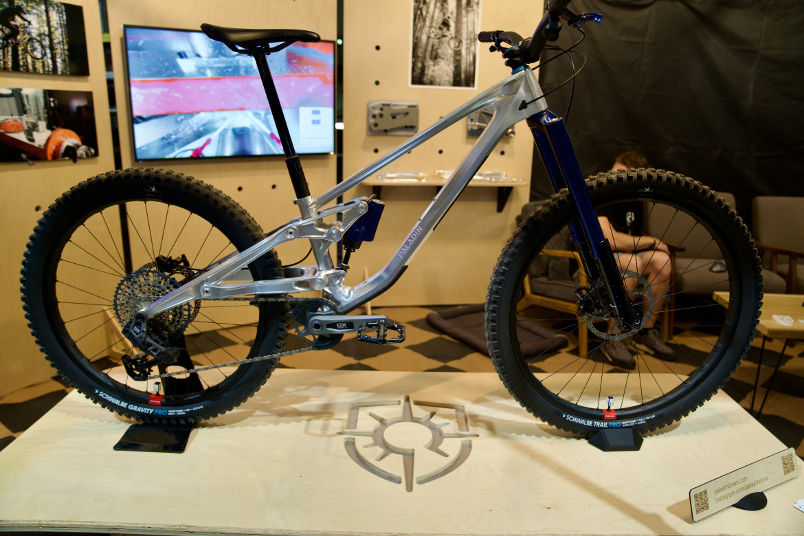 A modern mountain bike displayed on a wooden pedestal, showcasing its sleek aluminum frame and robust tires. The bike features a blue fork and multiple gears, with a detailed gear system visible at the rear wheel. In the background, there are photographs and a screen showing biking visuals, enhancing the bicycle's dynamic appeal. The setting includes wooden furniture and a relaxed atmosphere.