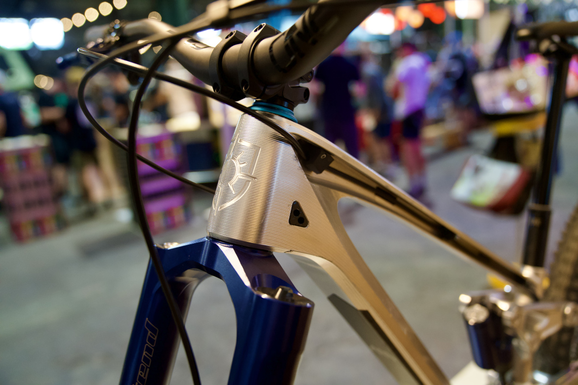 A close-up view of a modern mountain bike's frame, highlighting the sleek design and detailing on the head tube. The bike features a combination of metallic colors with a blue fork and a silver head tube engraved with a logo. The background shows a blurred scene of people interacting in a vibrant, market-like setting.