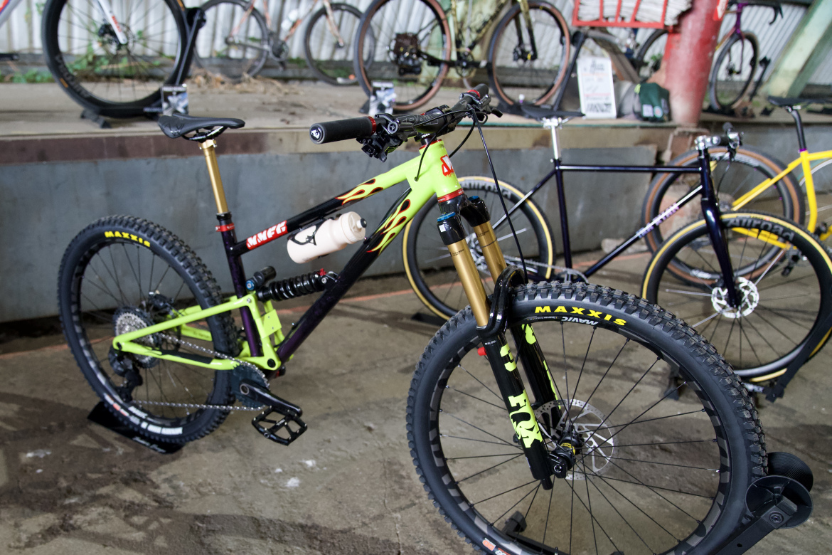 A brightly colored mountain bike with a green and black frame, featuring flame-like designs and a gold front fork. It has chunky tires with visible branding, a water bottle attached to the frame, and is positioned on a concrete floor among other bikes in a bike display area.