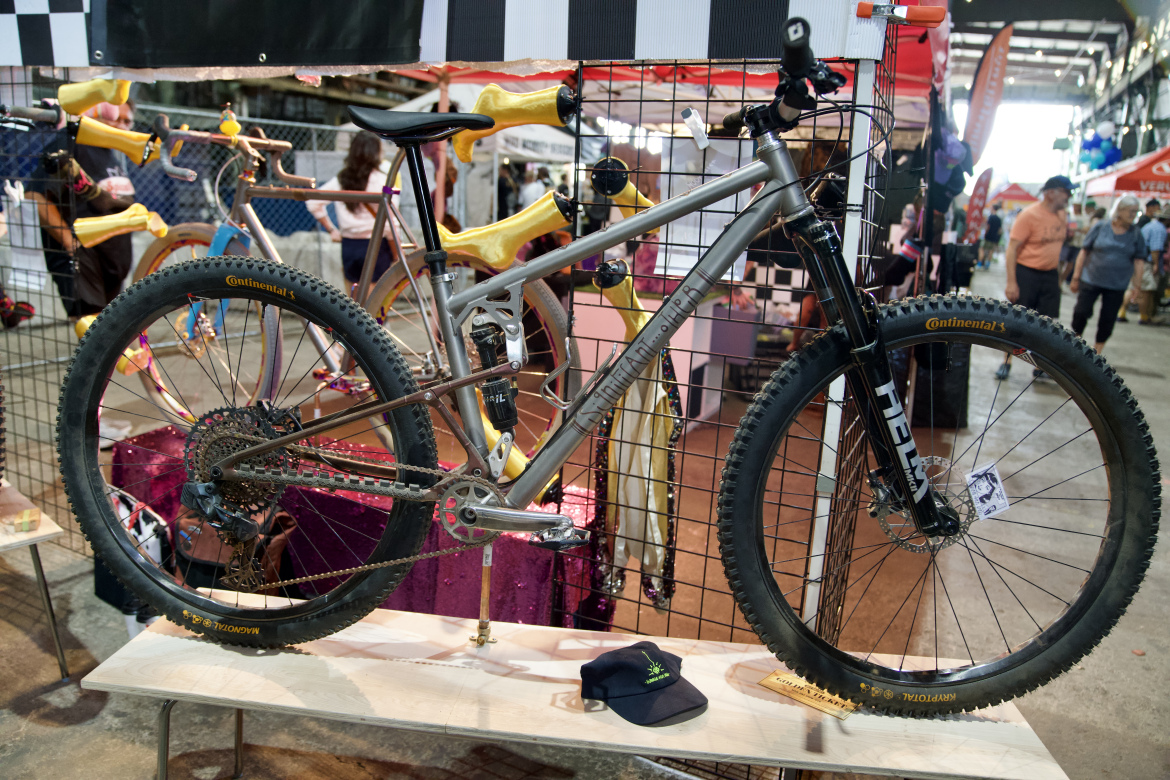 A silver mountain bike displayed on a wooden stand, featuring yellow handlebar grips and a Continental tire. In the foreground, a navy blue cap sits on the stand, while a colorful backdrop with hanging items and other bikes is visible behind the display. People can be seen moving through the area in the background.
