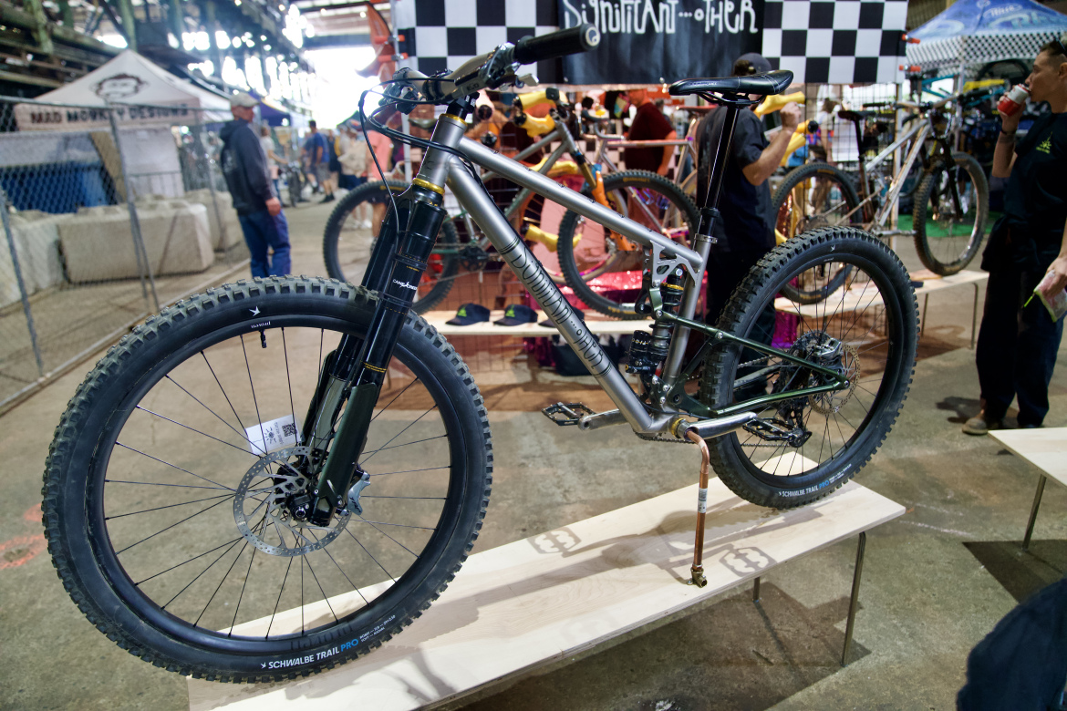 A mountain bike displayed on a wooden stand at a cycling event, featuring a metallic frame, thick tires, and visible suspension components. In the background, bicycles and people are seen in a bustling indoor setting, with various banners and booths.