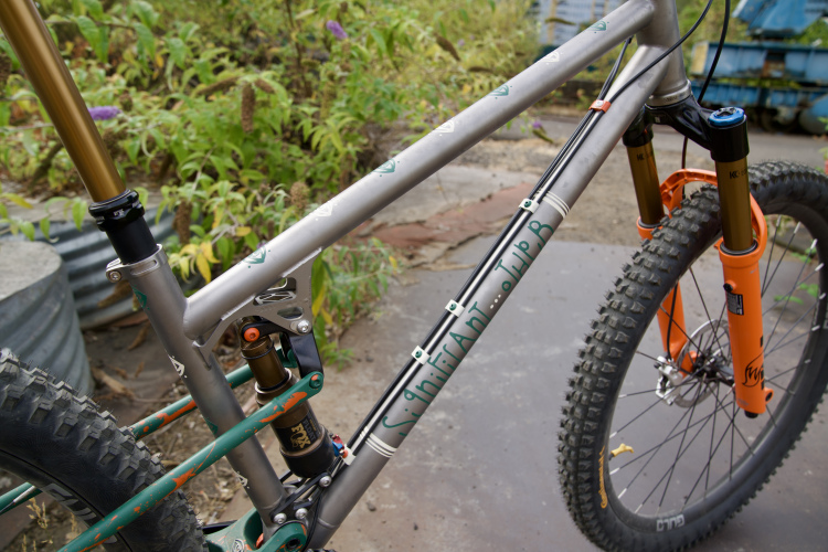 A close-up view of a mountain bike frame featuring a grey metallic finish with green and orange accents. The bike is equipped with a suspension system and has thick, textured tires. In the background, there are hints of foliage and some blurred objects, suggesting an outdoor setting.