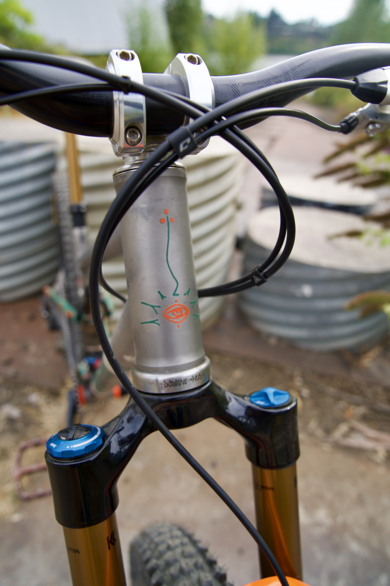 A close-up view of a bicycle's handlebars and frame, featuring a metallic fork with colorful graphics and cable routing. The handlebar grips are black, and there are blue knobs on the suspension fork. In the background, blurred corrugated metal containers and greenery can be seen, indicating an outdoor setting.