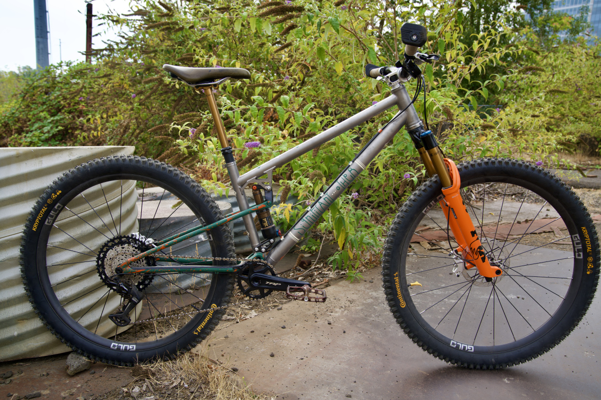 A mountain bike with a silver frame and orange front fork is positioned next to a metal container amidst greenery. The bike features thick, knobby tires suited for rugged terrain, and it has a rear suspension system. The background includes lush foliage, suggesting a natural outdoor setting.