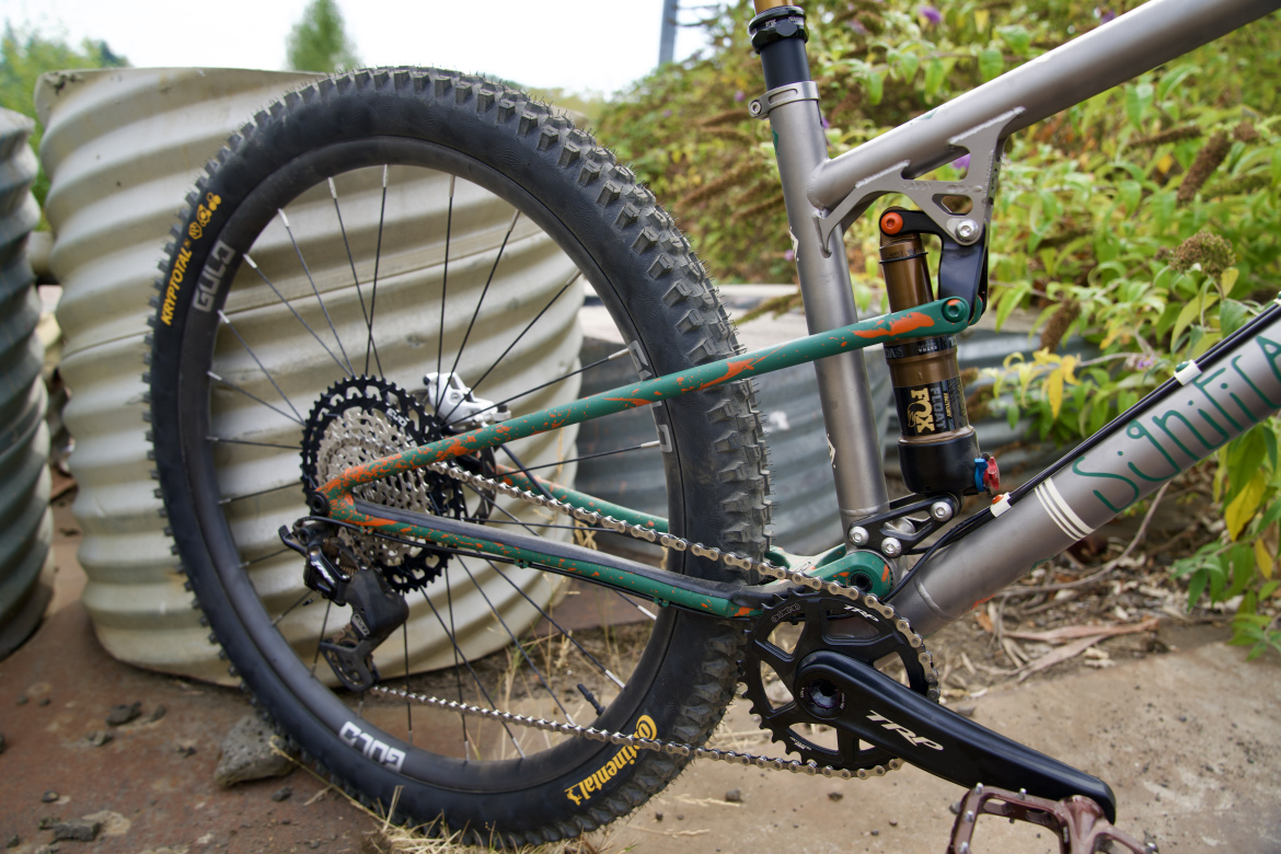 A close-up view of a mountain bike's rear wheel and frame, showcasing the suspension system, chain, and gears. The bike features a mix of green and silver colors with some visible wear and scratches. In the background, there are corrugated metal objects and greenery, indicating an outdoor environment.