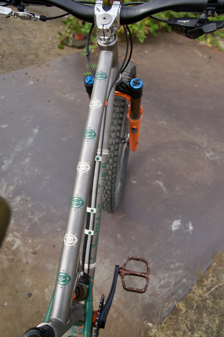 A close-up view of a mountain bike's aluminum frame, showcasing a unique design featuring green eye motifs along the top tube. The handlebars and front fork are visible, with black cables and decorative components. The bike is positioned on a rough surface near some greenery.