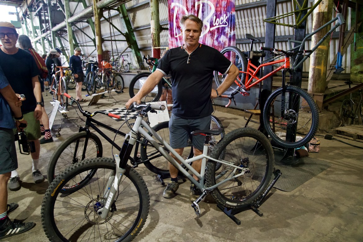 A man stands next to a gray mountain bike in an industrial setting filled with various bicycles. He is wearing a black shirt and gray shorts, with a relaxed expression. In the background, other individuals are mingling around more bikes, showcasing a vibrant atmosphere of a bike event. A colorful banner is partially visible on the wall.