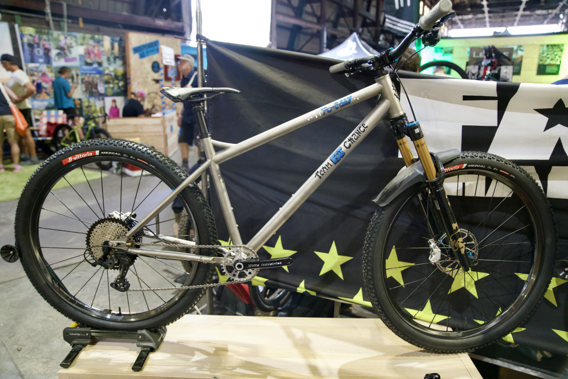 A high-performance mountain bike with a silver frame is displayed on a wooden stand. The bike features thick, knobby tires labeled "Vittoria Mezcal," a front suspension fork, and a modern drivetrain. In the background, a trade show setting can be seen, with various people and colorful banners.
