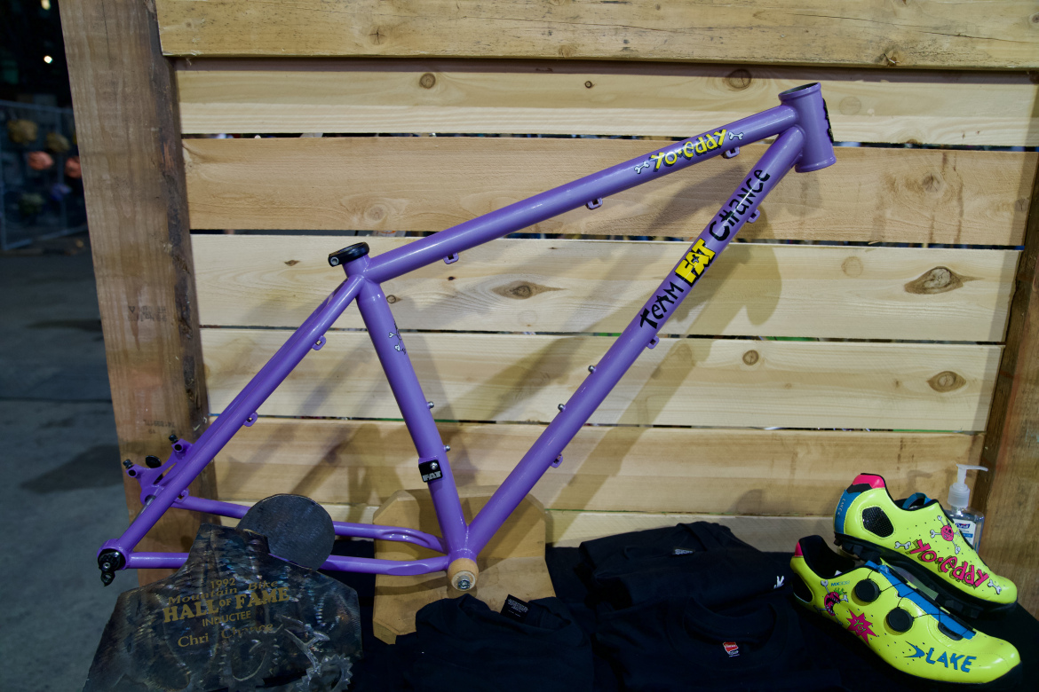 A purple bicycle frame displaying colorful graphics is positioned against a wooden backdrop. In front of the frame, there are black t-shirts neatly arranged and a pair of bright yellow cycling shoes with pink accents. A small trophy engraved with "Mountain Bike Hall of Fame" is also visible on the display.