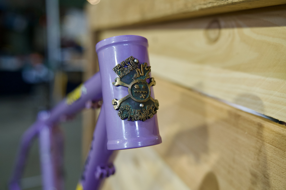 A close-up view of a purple bicycle frame showcasing a decorative emblem featuring a skull and crossbones design, set against a wooden background.