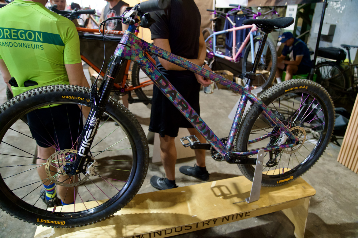 A colorful mountain bike displayed on a wooden stand, with the word "DARWIN" boldly printed on its frame. A rider in a green cycling jersey stands nearby, while other bikes are visible in the background, indicating an event or exhibition. The setting appears to be indoors, with dim lighting and a casual atmosphere.