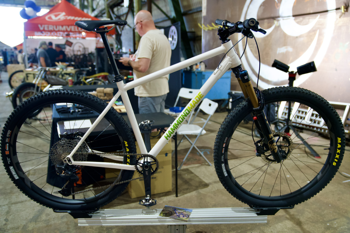 A white mountain bike with a distinctive green logo stands on a display. The bike features thick tires and a front suspension fork, showcasing its off-road capability. In the background, a blur of people and a red booth can be seen, indicating a bike show or event setting.