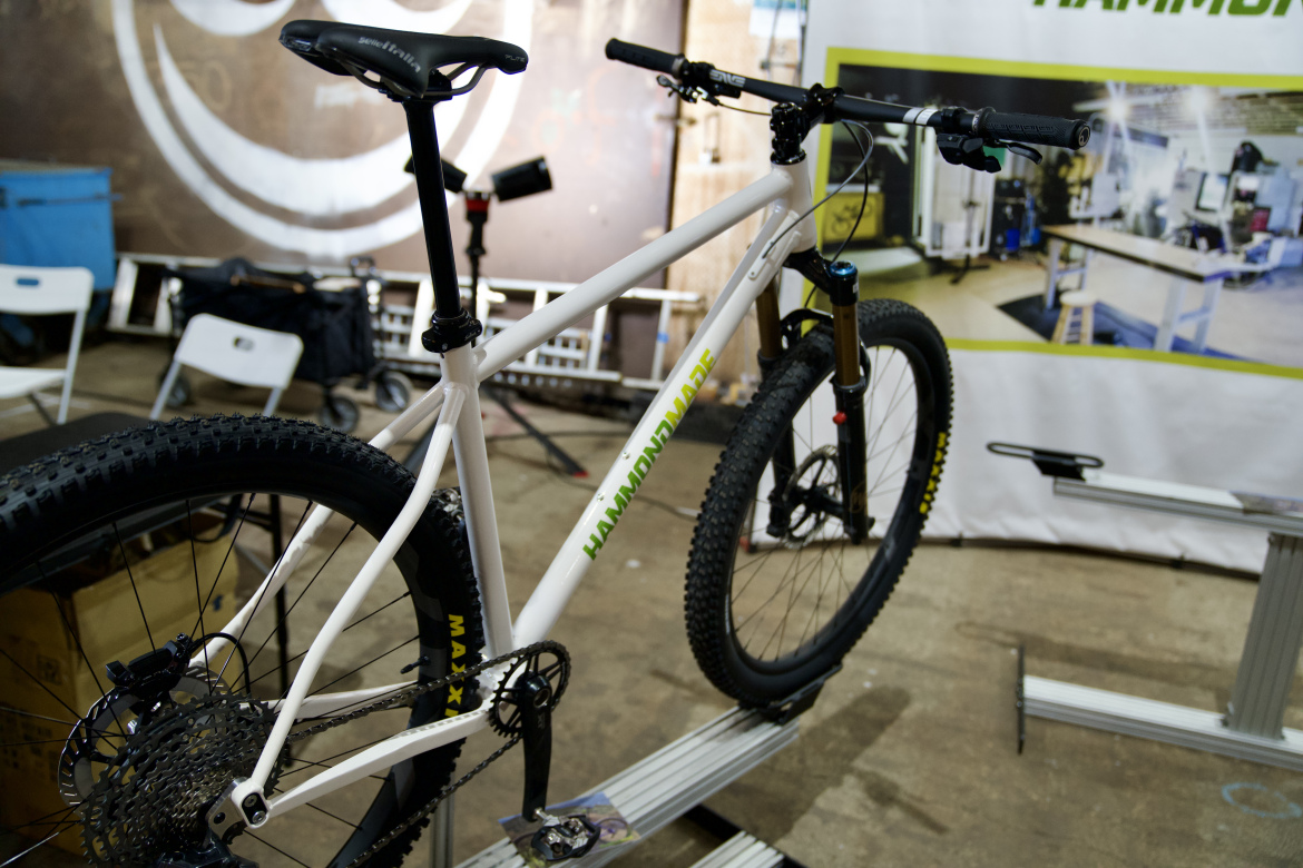 A close-up view of a mountain bike featuring a white frame with green accents, mounted on a display. The bike has thick, rugged tires suitable for off-road terrain, a black seat, and visible components like a derailleur and brakes. In the background, a partially blurred exhibition area with a banner displaying the brand name can be seen, along with some workshop equipment and seating.