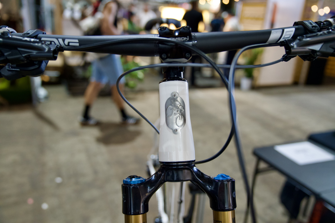 Close-up view of a mountain bike's handlebars and fork, featuring a prominent white head tube with a decorative emblem. The bike has black and gold components, with cables running neatly along the frame. In the blurred background, people can be seen in a bustling indoor environment.