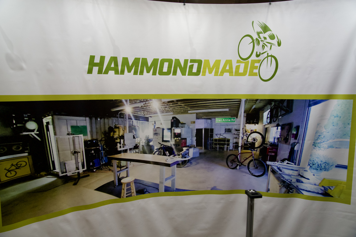 A banner featuring the word "HAMMONDMADE" in bold green letters at the top, above a panoramic image of a workshop. The workshop is well-lit and contains various tools and equipment, including a workbench with a stool, bikes, and machinery arranged along the walls, with a street sign reading "Glen Annie Rd" visible in the background.