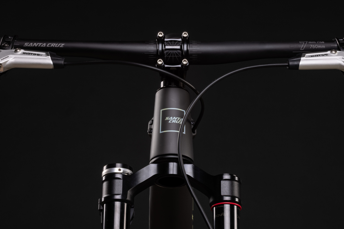 Alt text: Close-up view of a mountain bike's handlebars and front suspension, featuring a black Santa Cruz brand handlebar, control levers, and a matte black fork.
