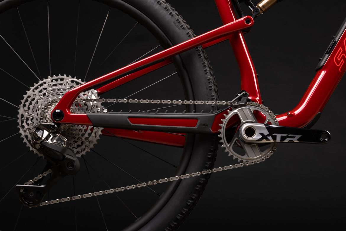 Close-up view of the drivetrain of a mountain bike featuring a red frame, with visible cassette, chain, and crankset components. The image highlights the intricate details of the bike's gearing system against a dark background.