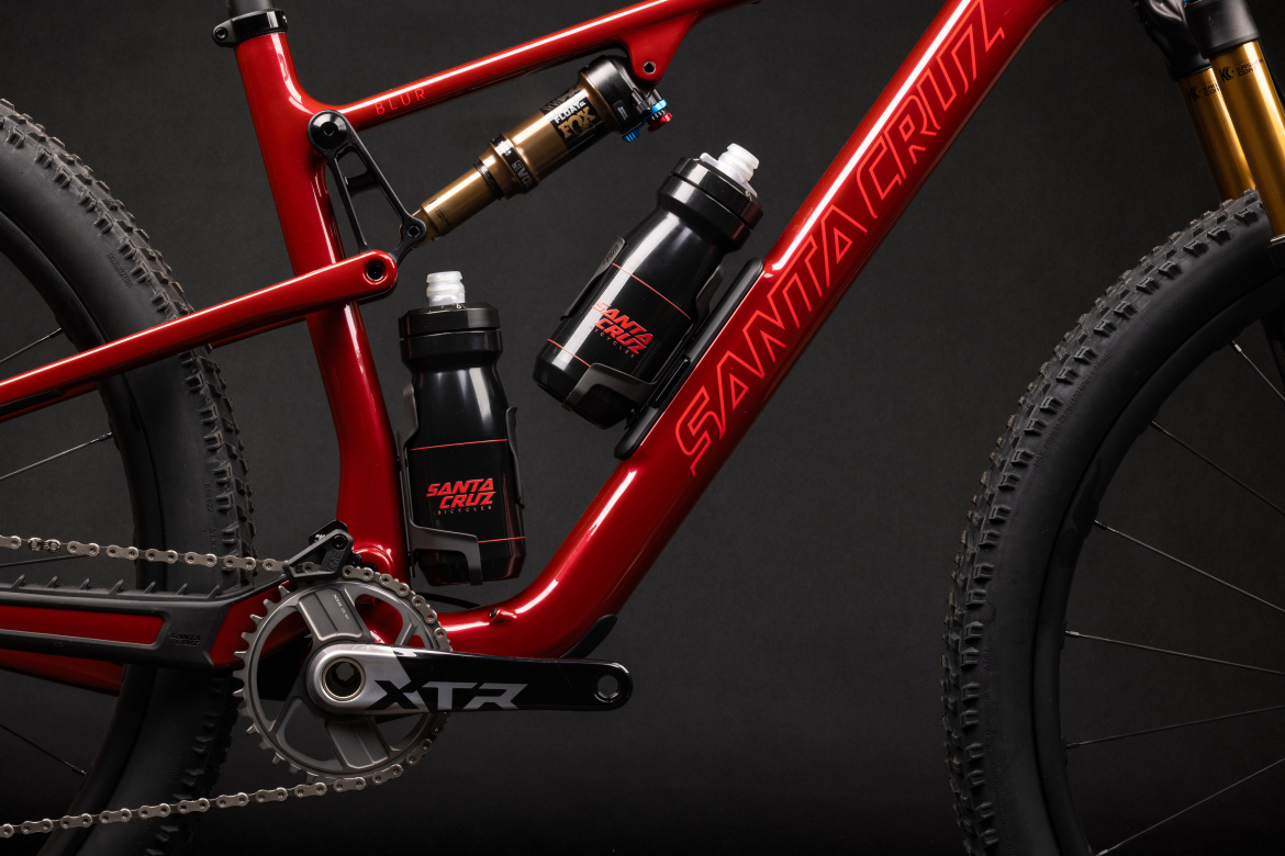 A close-up view of a sleek red mountain bike frame featuring the brand name "Santa Cruz." The bike has a dual suspension setup with a shock absorber visible. Two black water bottles are mounted on the frame, and the bike's drivetrain includes a chainring labeled "XTR." The background is dark, enhancing the bike's vibrant color and details.