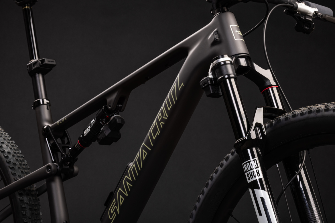 Close-up image of a modern mountain bike featuring a sleek black frame with the "Santa Cruz" logo prominently displayed. The bike includes a RockShox fork and a shock absorber with a compact design, emphasizing its performance capabilities. The background is dark, highlighting the bike's details and craftsmanship.