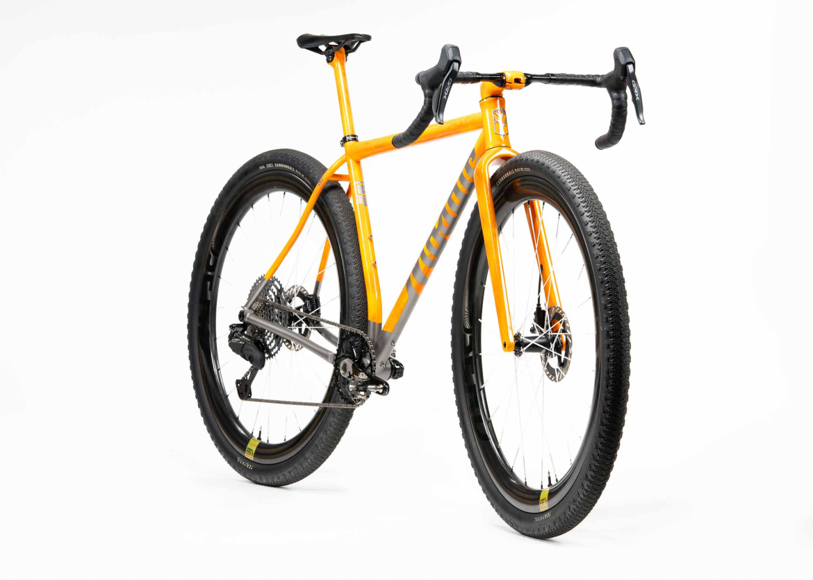 A bright orange and gray gravel bike with wide tires, featuring a sleek frame and a drop handlebar design. The bike is positioned at an angle against a white background, showcasing its components, including a rear derailleur and disc brakes.