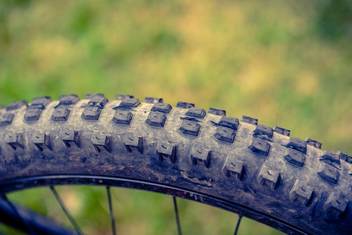Close-up view of a mountain bike tire showing the tread pattern, which features knobby rubber for improved traction on rough terrain, with a blurred green background.