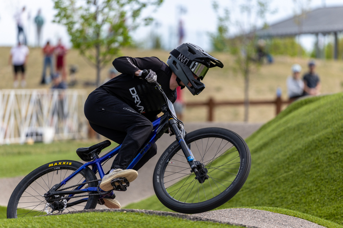 A cyclist in a black helmet and gear leans into a turn on a blue mountain bike while navigating a course with grassy hills and a dirt path. Spectators are visible in the background.