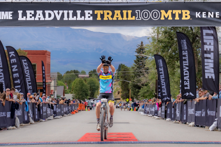 A mountain biker crossing the finish line at the Leadville Trail 100 MTB race, raising both hands in celebration amidst a cheering crowd. Colorful flags and a scenic backdrop of mountains and trees frame the event, capturing the excitement of the moment.