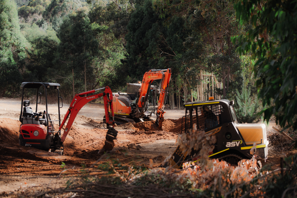 A construction site featuring three pieces of heavy machinery working on digging and earthmoving. A red mini excavator is in the foreground, actively digging into the ground. An orange excavator is in the middle distance, also engaged in excavation, while a small black and yellow skid steer loader can be seen to the right, maneuvering through the site. Surrounding the area are trees and foliage, creating a natural backdrop for the construction work.