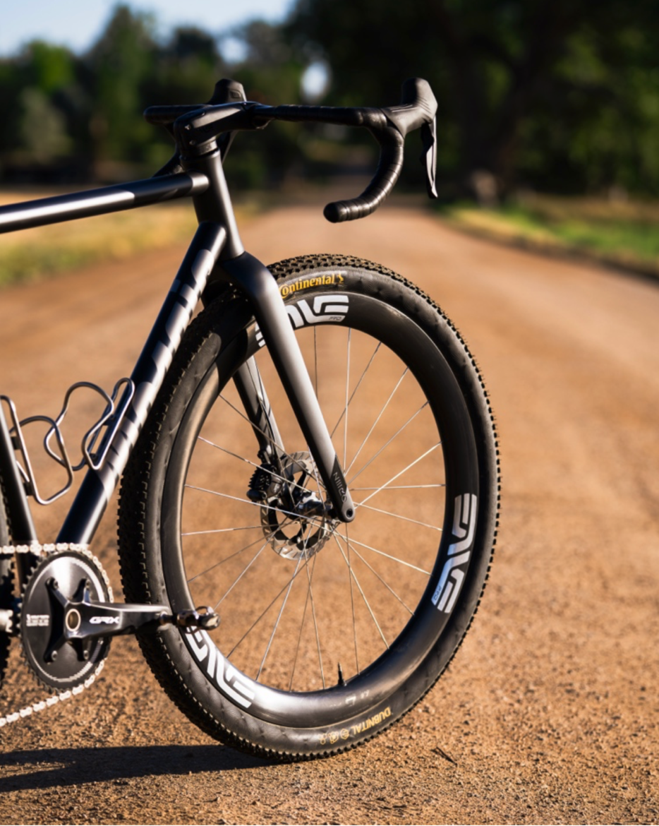 Close-up of a black gravel bike positioned on a dirt path, showcasing the rear wheel, disc brake, and part of the bike's frame and handlebars, with a blurred natural landscape in the background.