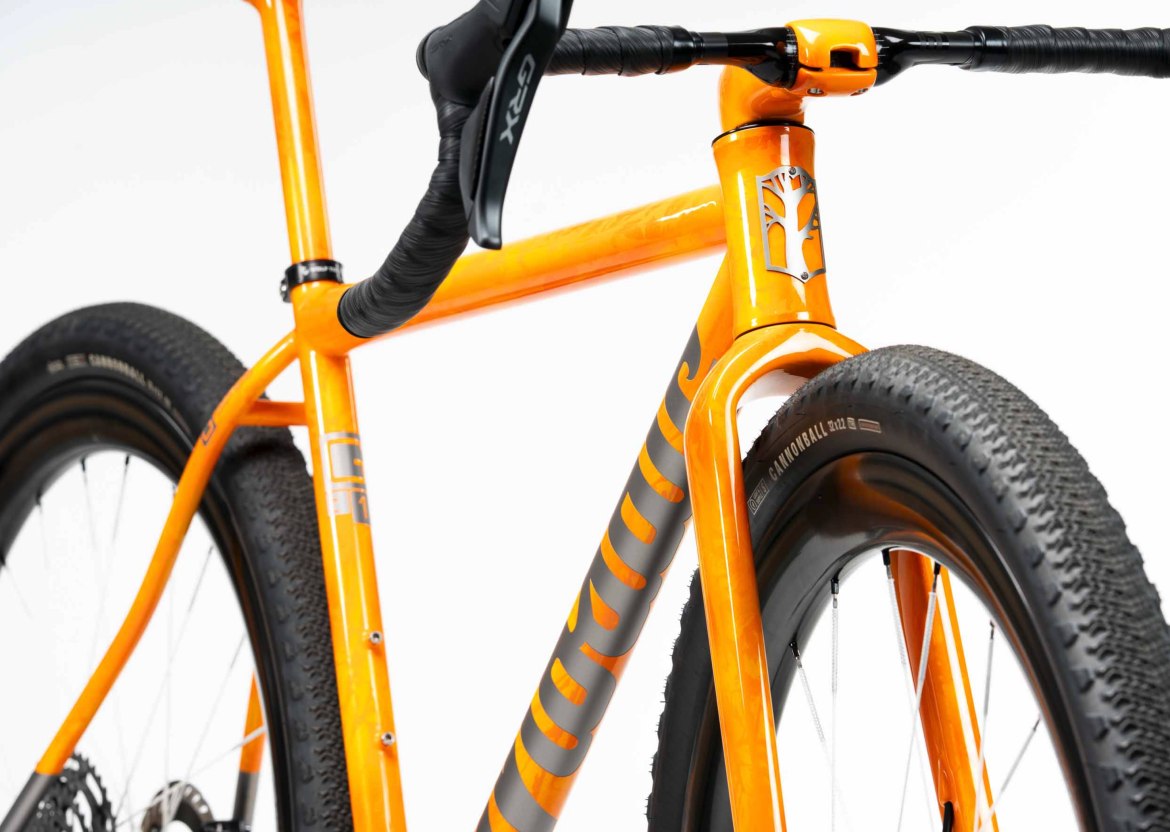 Close-up view of an orange bicycle frame, showcasing the intricate design and details, including the handlebars and textured tires. The image highlights the unique color and craftsmanship of the bike, set against a clean white background.