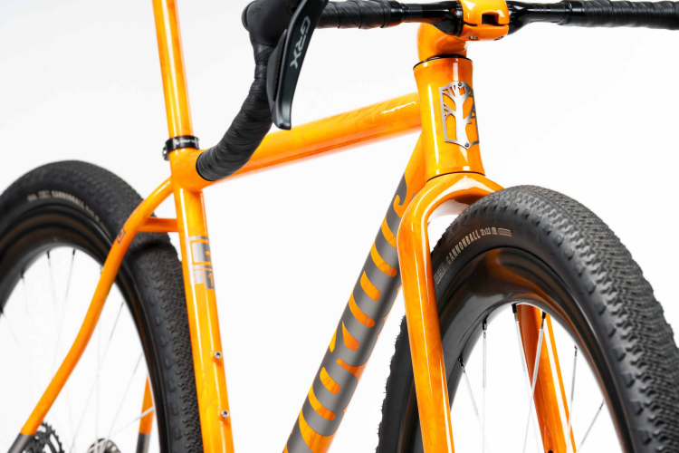 Close-up view of an orange bicycle frame, showcasing the intricate design and details, including the handlebars and textured tires. The image highlights the unique color and craftsmanship of the bike, set against a clean white background.